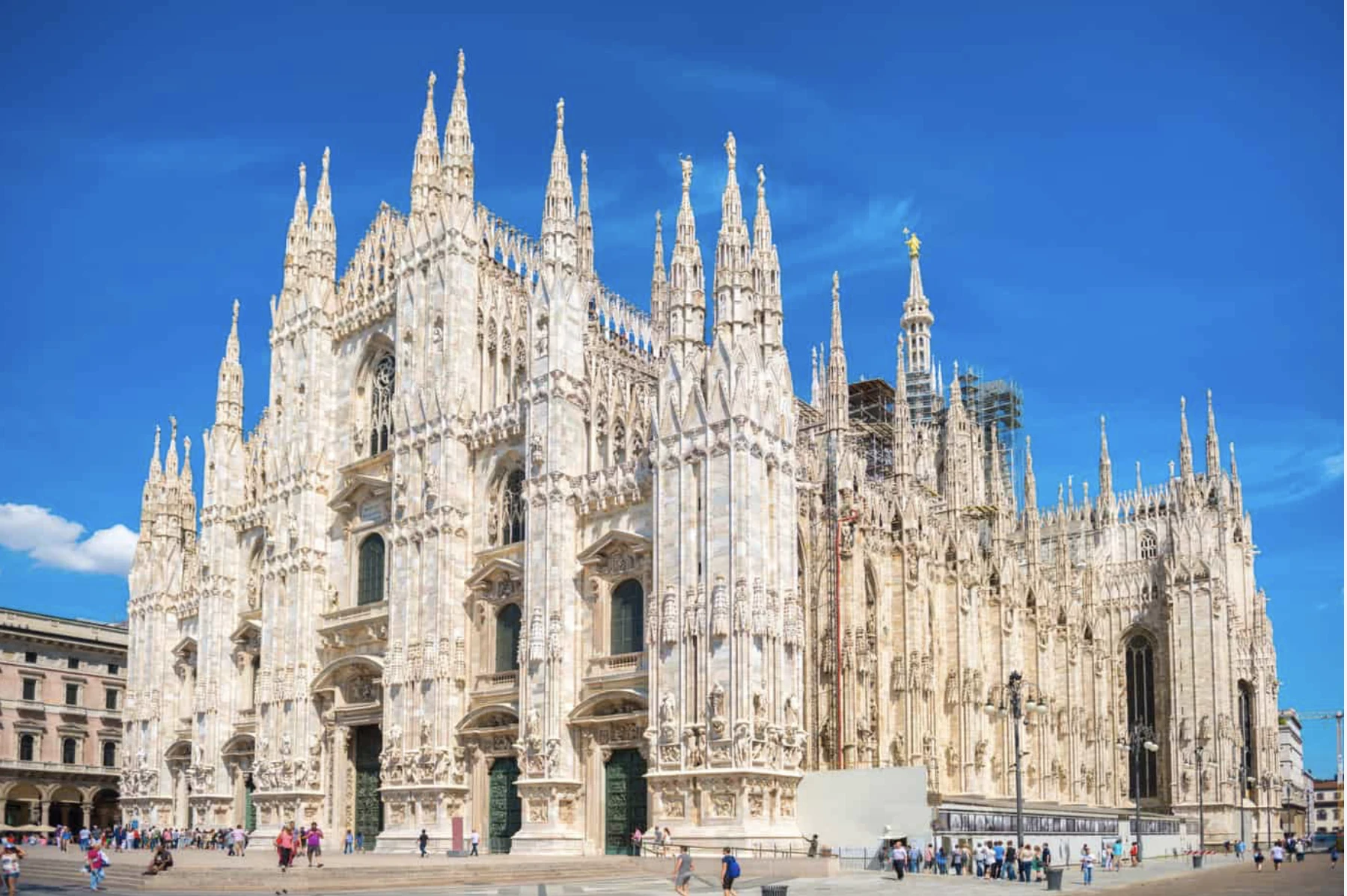 Fachada y agujas de mármol del Duomo de Milán bajo un cielo azul intenso, con visitantes en la plaza.