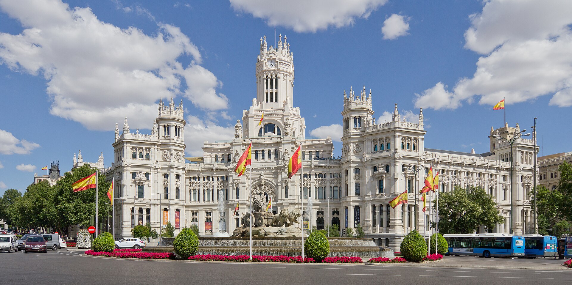 Palacio de Cibeles bajo un cielo azul con nubes, fuente, flores, árboles y actividad urbana.